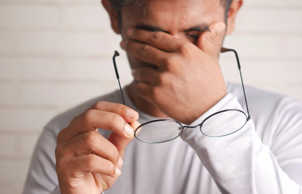 Person holding a pair of glasses and rubbing their eyes as if experiencing discomfort.