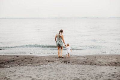 View of the back of a mother and her child holding hands on a beach as they stand at the edge of the water