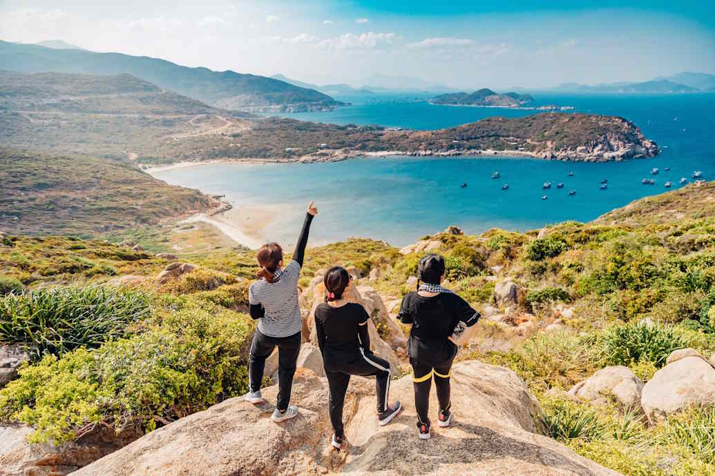 Three people stood on top of a cliff, one pointing to the sky, that overlooks a blue expanse of water with islands.