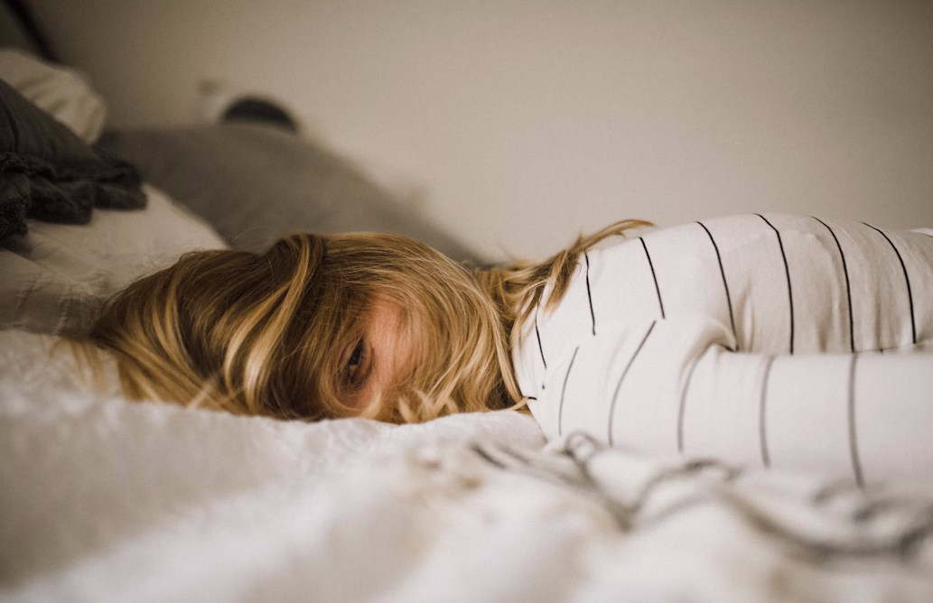 Woman in striped top lying on her front on a bed with her face turned to the side and her eyes looking at the camera through her hair.